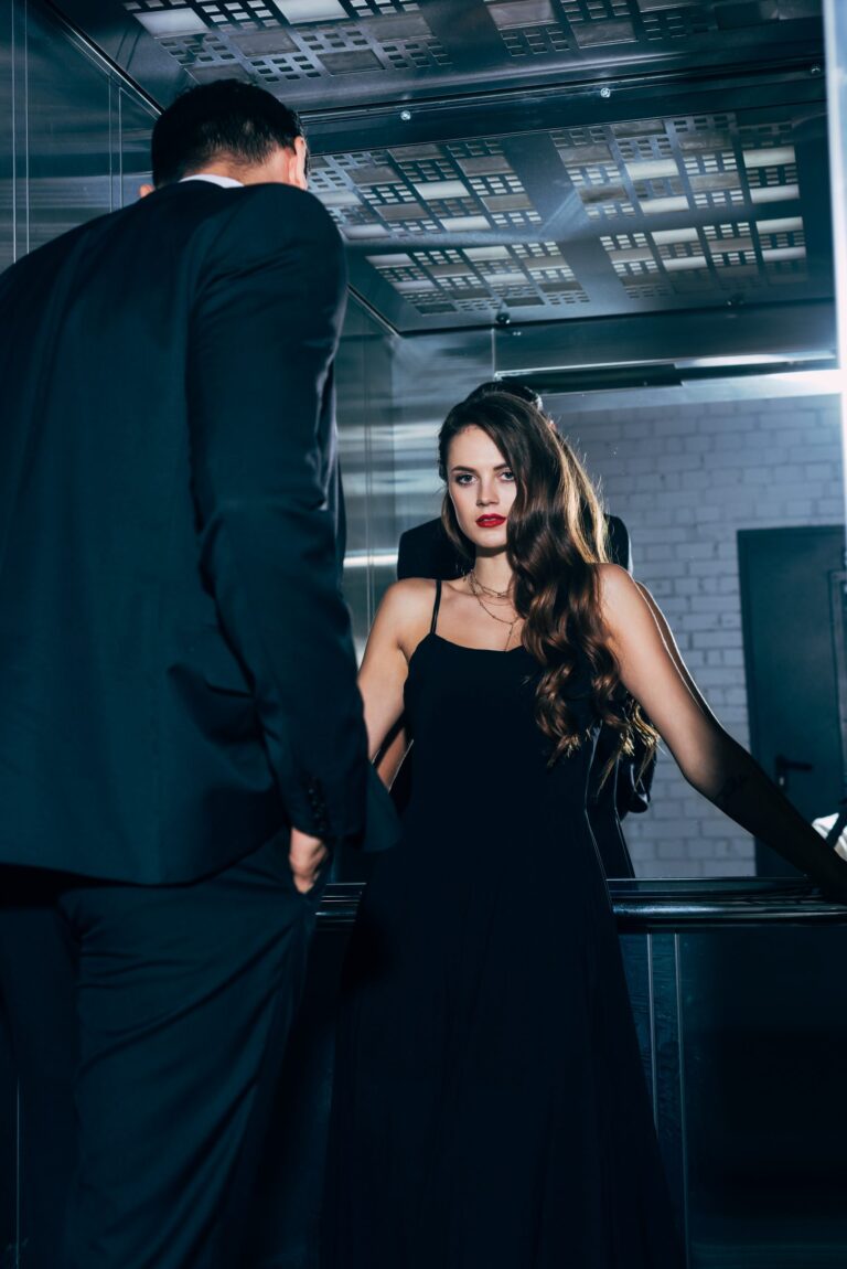 beautiful seductive woman in black dress looking at camera with man on foreground in elevator